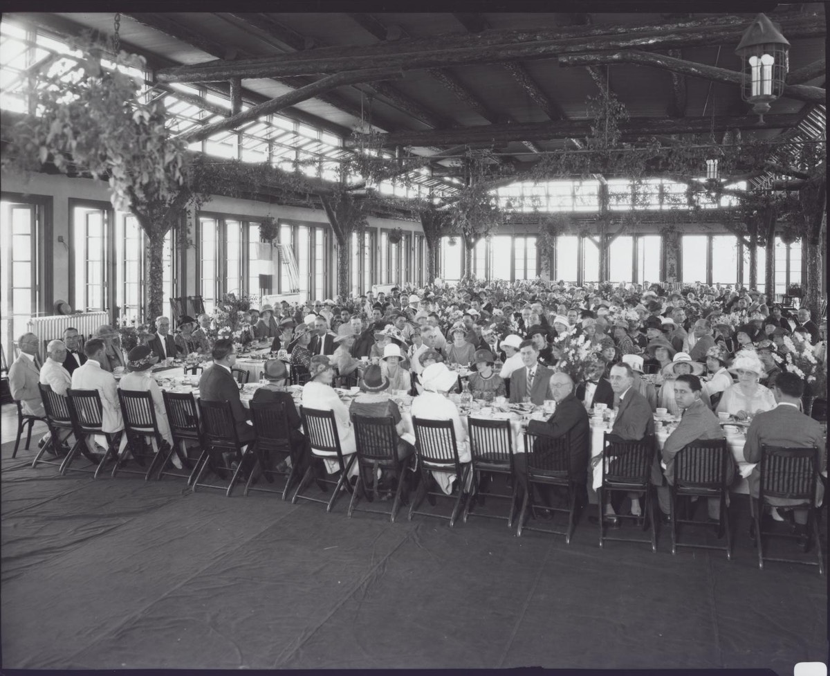 Photo of many men in suits and women in hats seated at long tables with white tablecloths under an open-air terrace roof